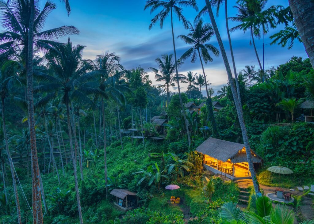 A serene tropical hut illuminated at twilight in a lush Balinese forest setting, surrounded by tall palm trees.